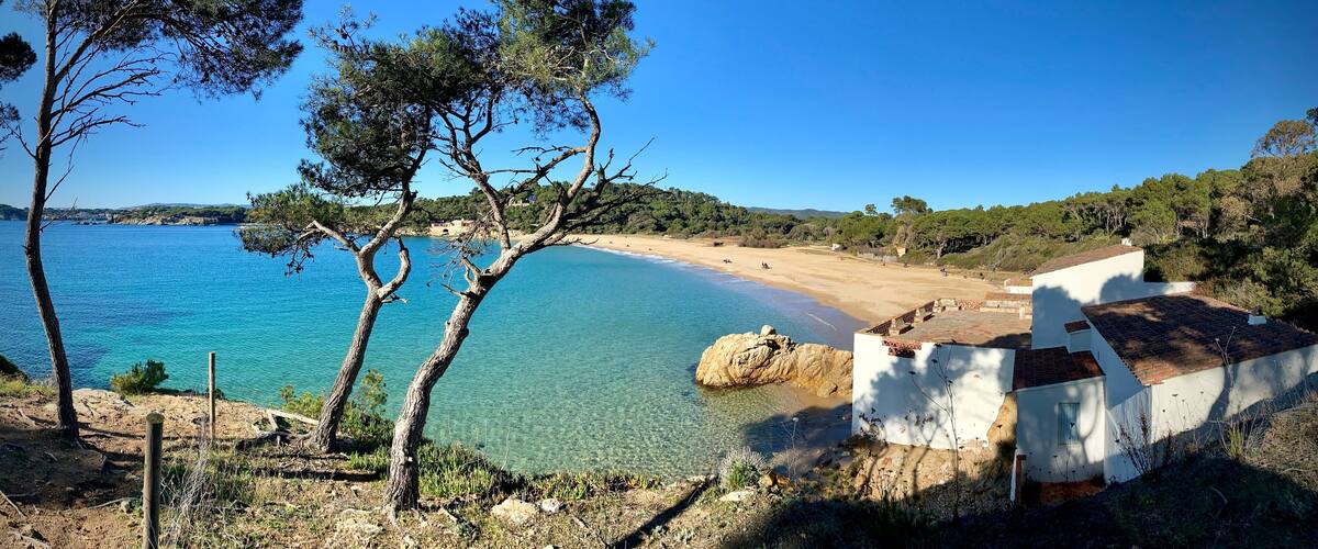 A coastal landscape with clear blue waters, a sandy beach, and a white building with a tiled roof.