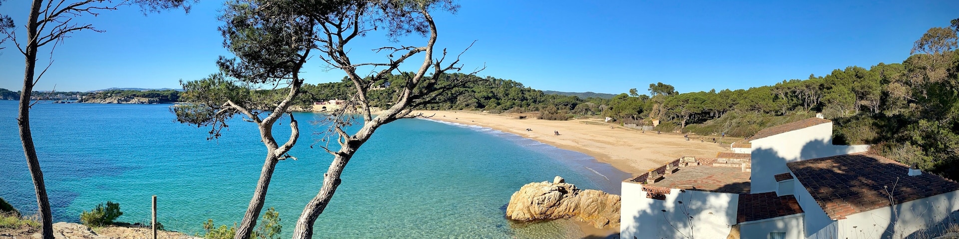 A coastal landscape with clear blue waters, a sandy beach, and a white building with a tiled roof.