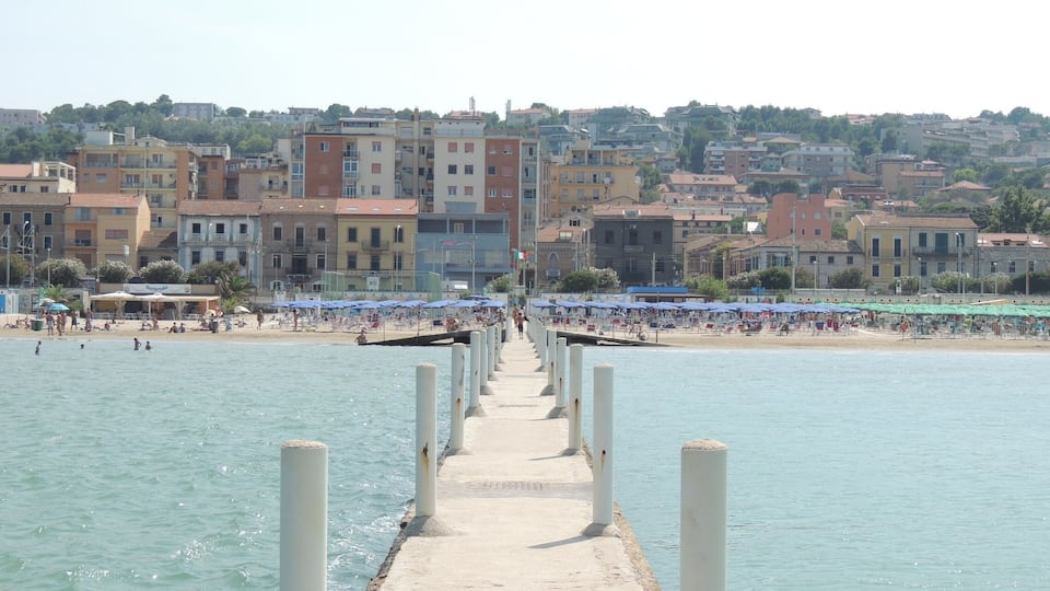 A pier extending into calm waters with a beach and buildings in the background.