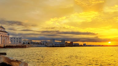Paisagem costeira urbana com um edifício histórico imponente, um calçadão à beira-mar e um pôr do sol sobre o oceano.