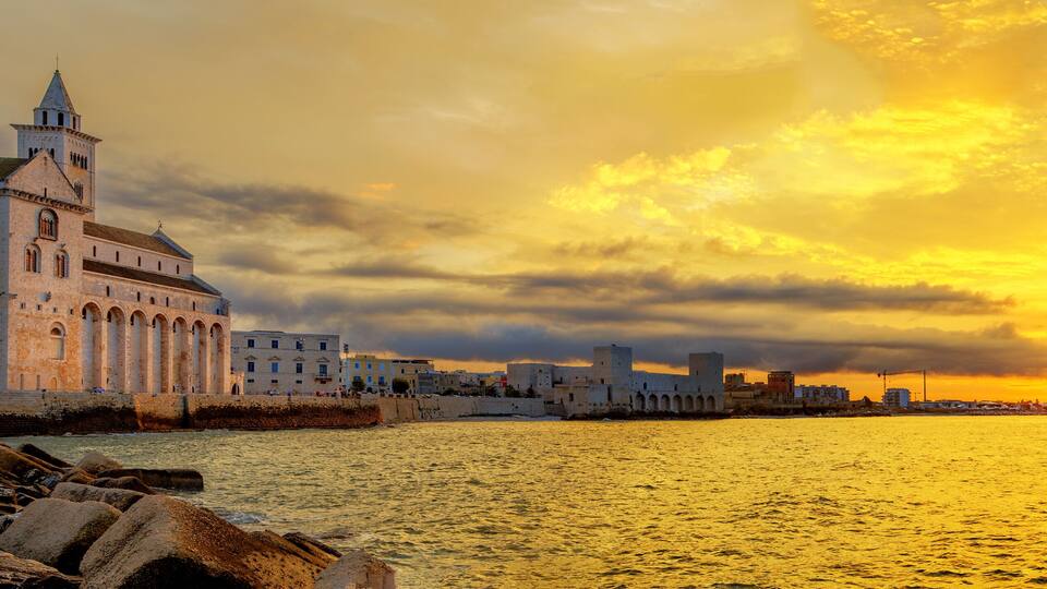 A coastal cityscape with a prominent historical building, a waterfront promenade, and a sunset over the sea.
