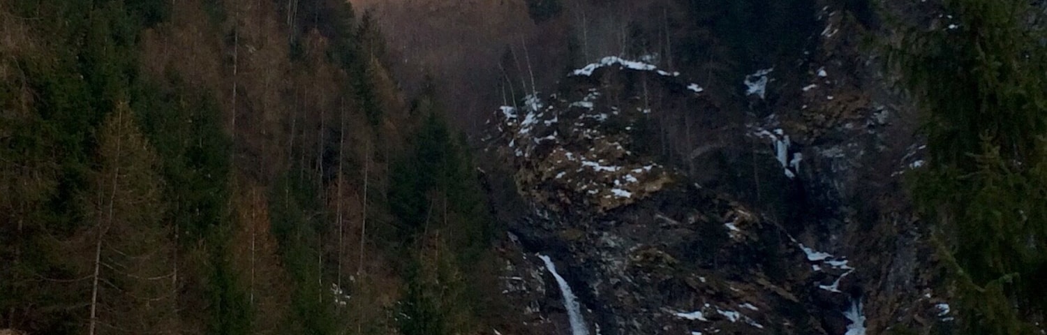 Paisaje montañoso con cascada, suelo cubierto de nieve y denso bosque.