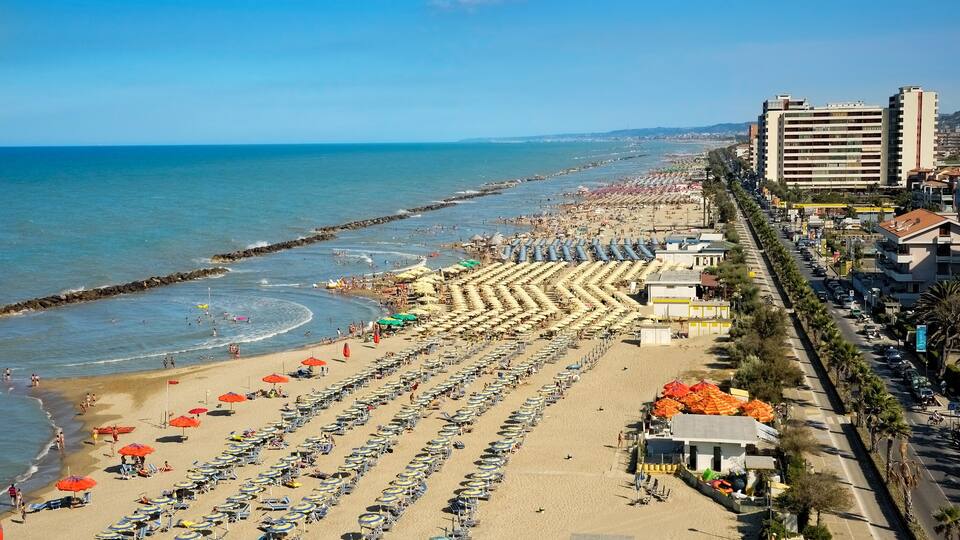 A beach with rows of umbrellas and sunbeds, a promenade with buildings, and a clear blue sky.