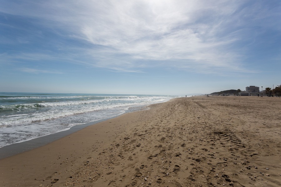 A sandy beach with footprints, waves, and buildings in the background.