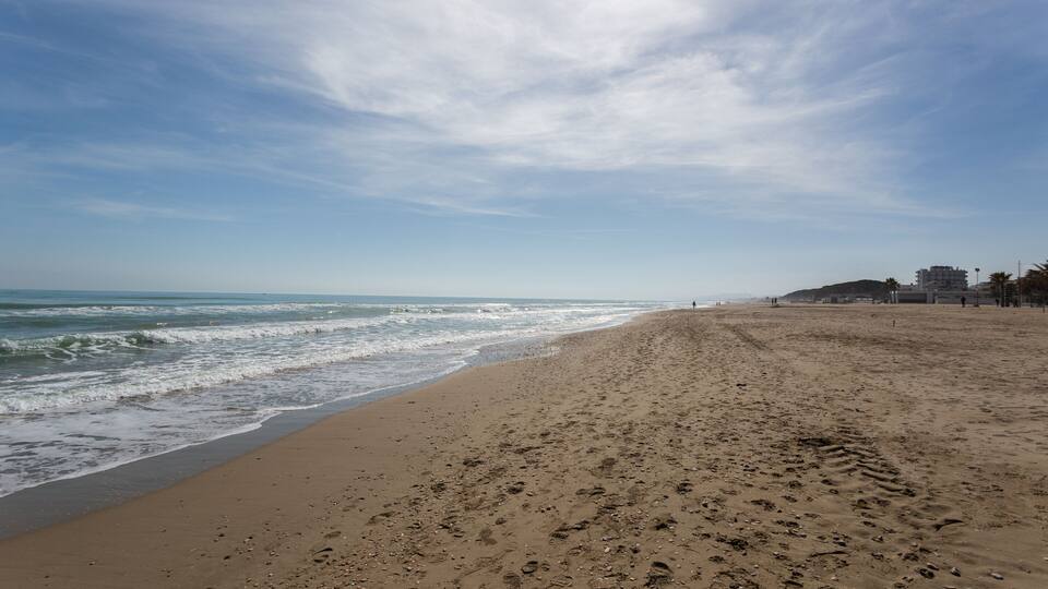 A sandy beach with footprints, waves, and buildings in the background.