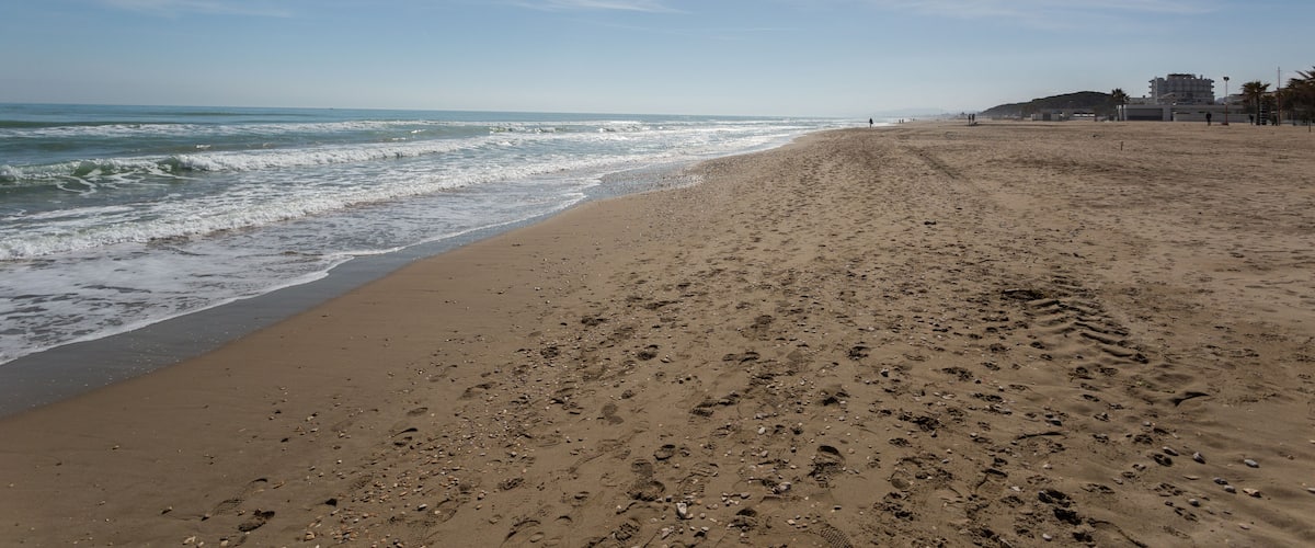 A sandy beach with footprints, waves, and buildings in the background.