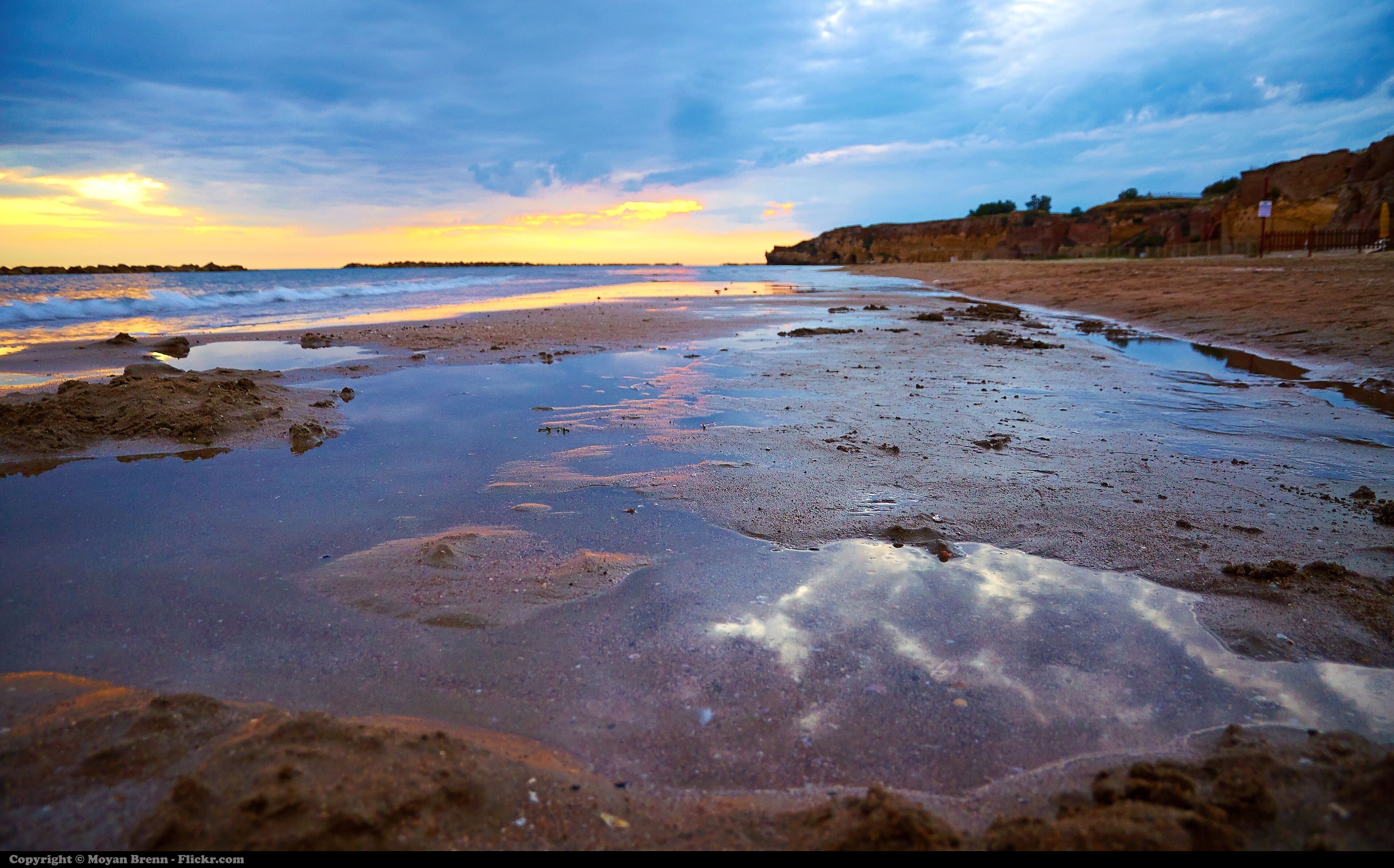 A beach with wet sand and puddles, a rocky cliff, and a sunset sky.