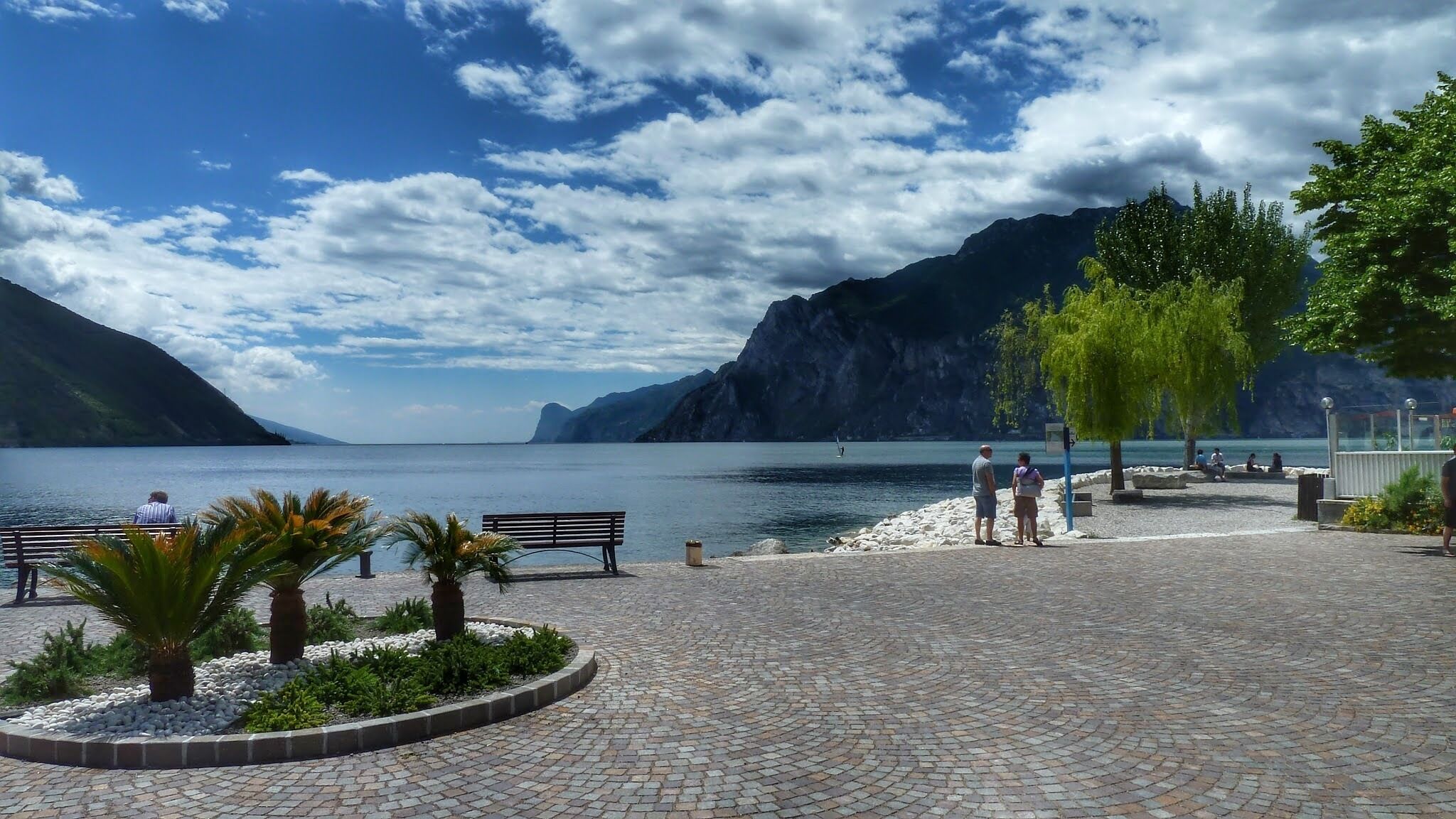 A paved waterfront promenade with benches, palm trees, and a view of a lake and mountains.