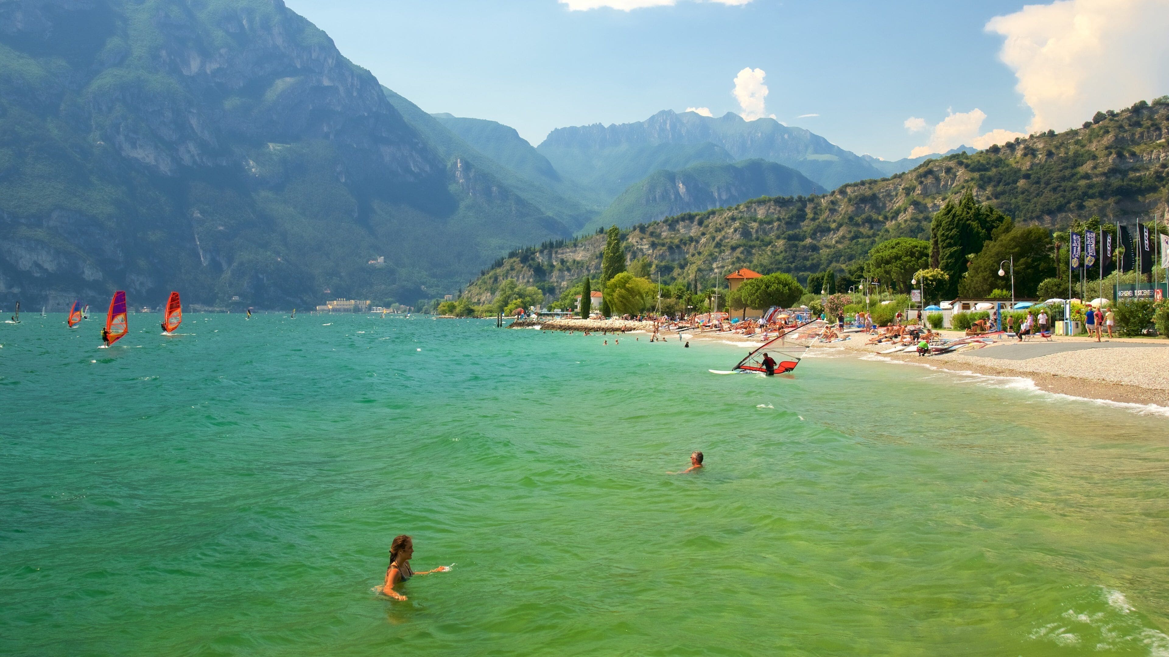 A beach with clear turquoise water, people swimming and windsurfing, and mountains in the background.
