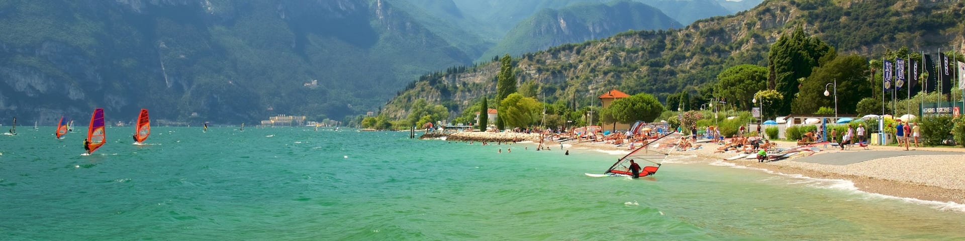 A beach with clear turquoise water, people swimming and windsurfing, and mountains in the background.