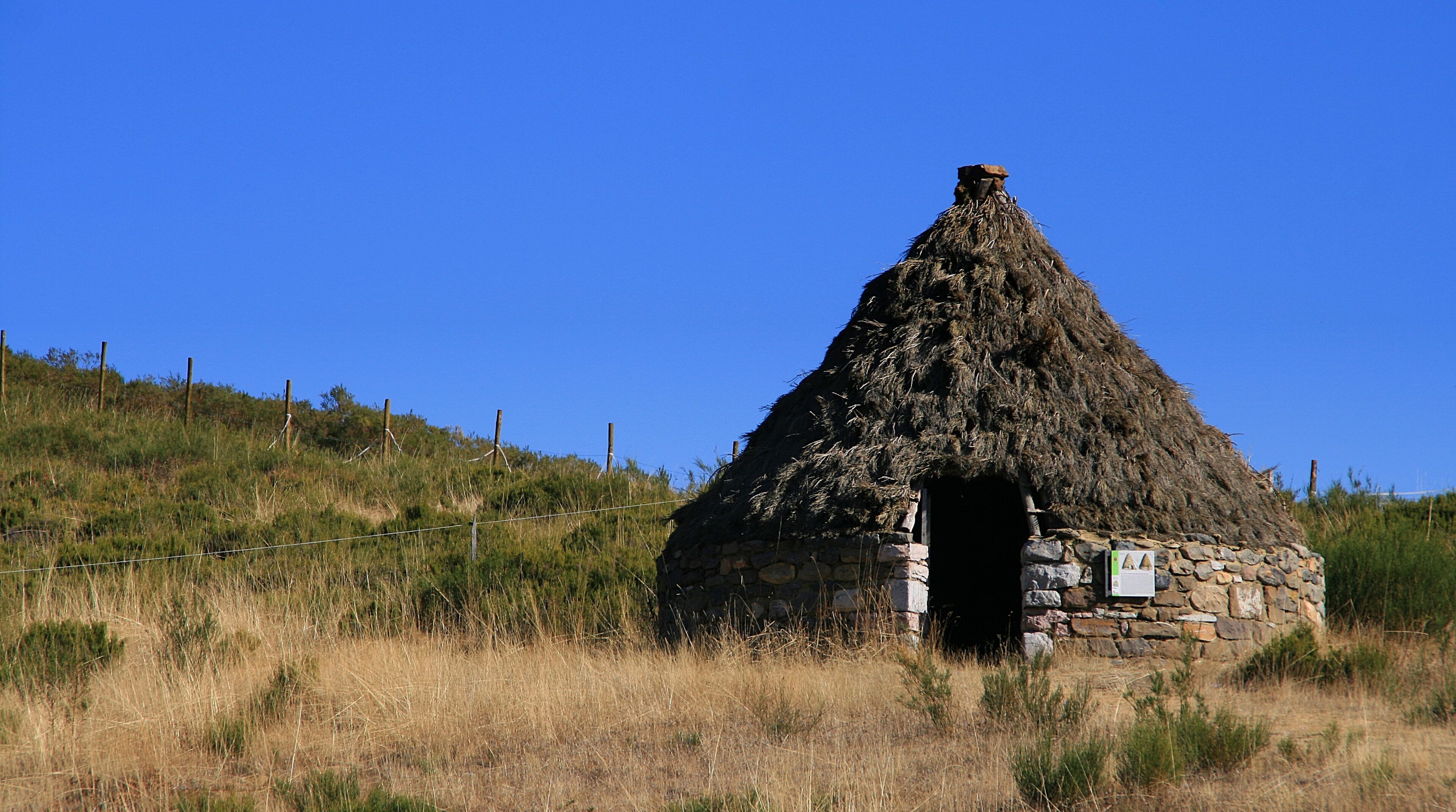 A stone hut with a thatched roof, surrounded by dry grass and a clear blue sky.