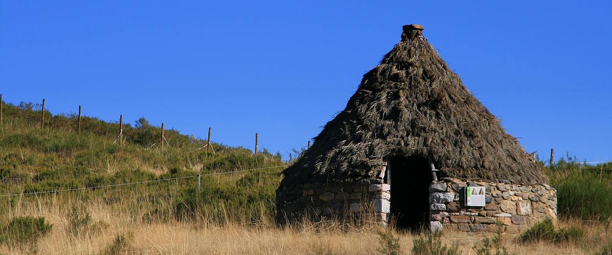 A stone hut with a thatched roof, surrounded by dry grass and a clear blue sky.