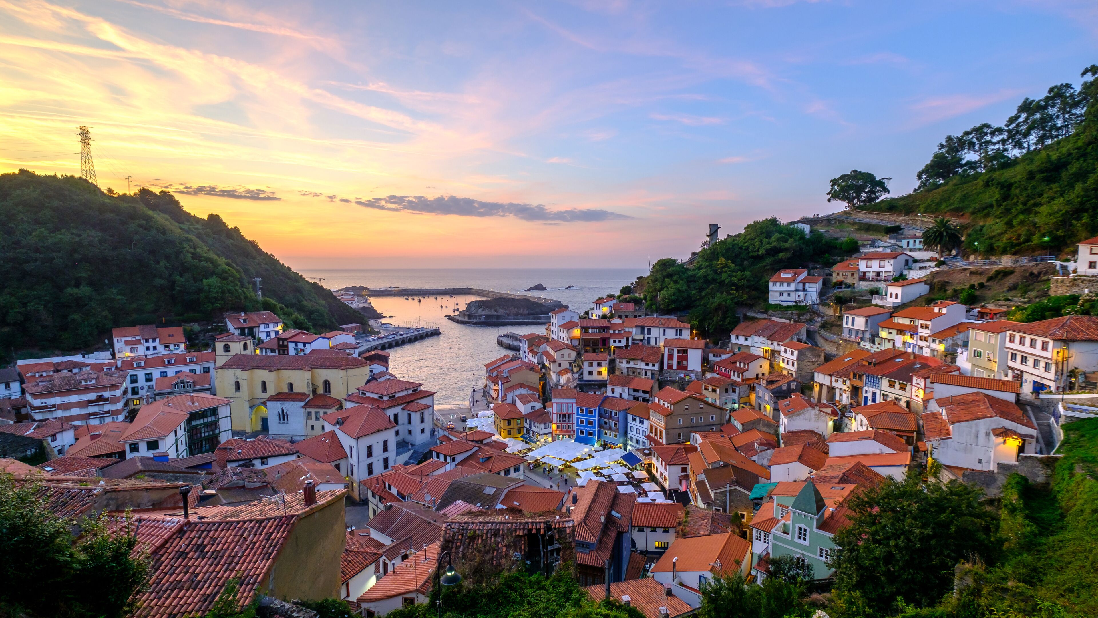 A coastal town with terracotta-roofed buildings, a pier extending into the sea, and a sunset sky.