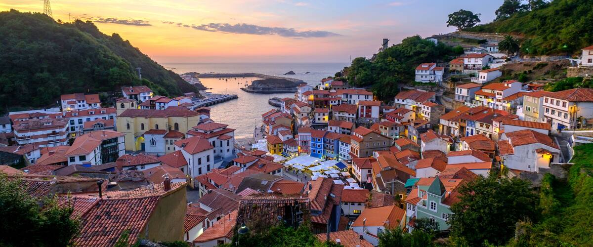 A coastal town with terracotta-roofed buildings, a pier extending into the sea, and a sunset sky.