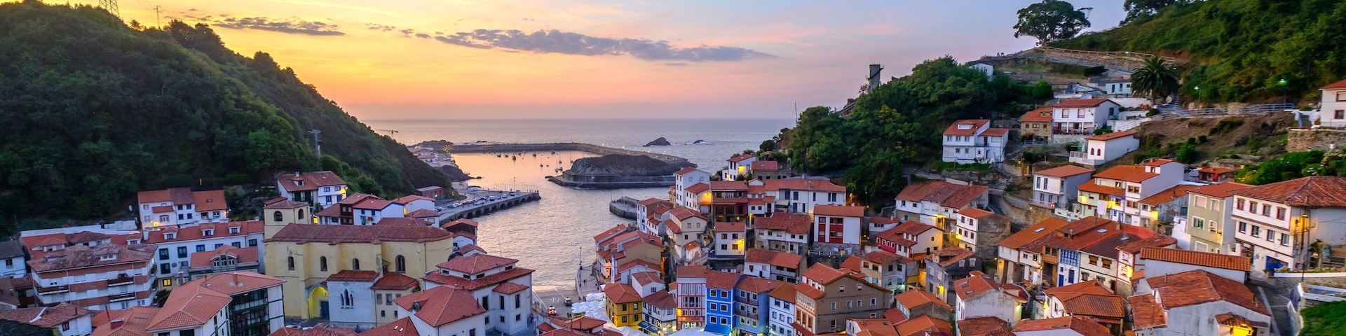 A coastal town with terracotta-roofed buildings, a pier extending into the sea, and a sunset sky.