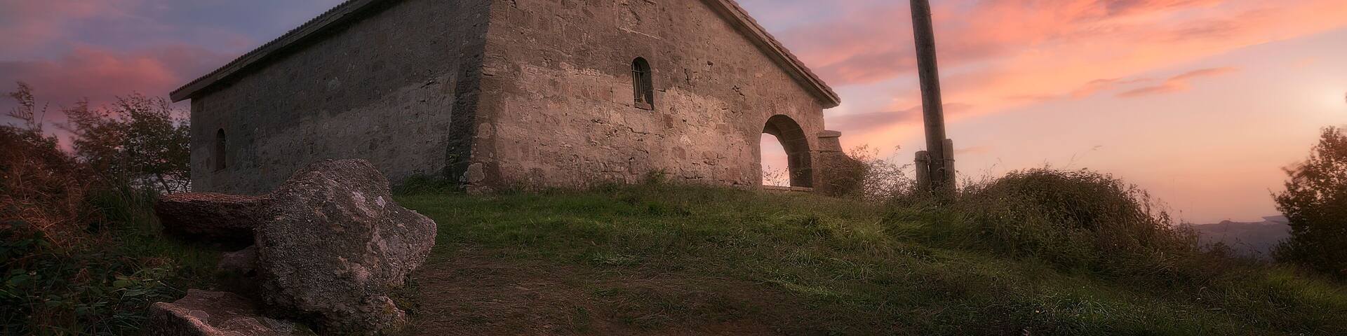 Hermitage of Santa Cruz at the top of Mount Bizkargi in Amorebieta, Bizkaia, with the ikurriña waving, it features a dramatic sky with warm clouds at sunset