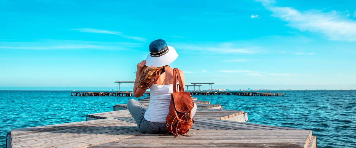 A woman sitting on a wooden pier with a clear blue sky and calm sea in the background.