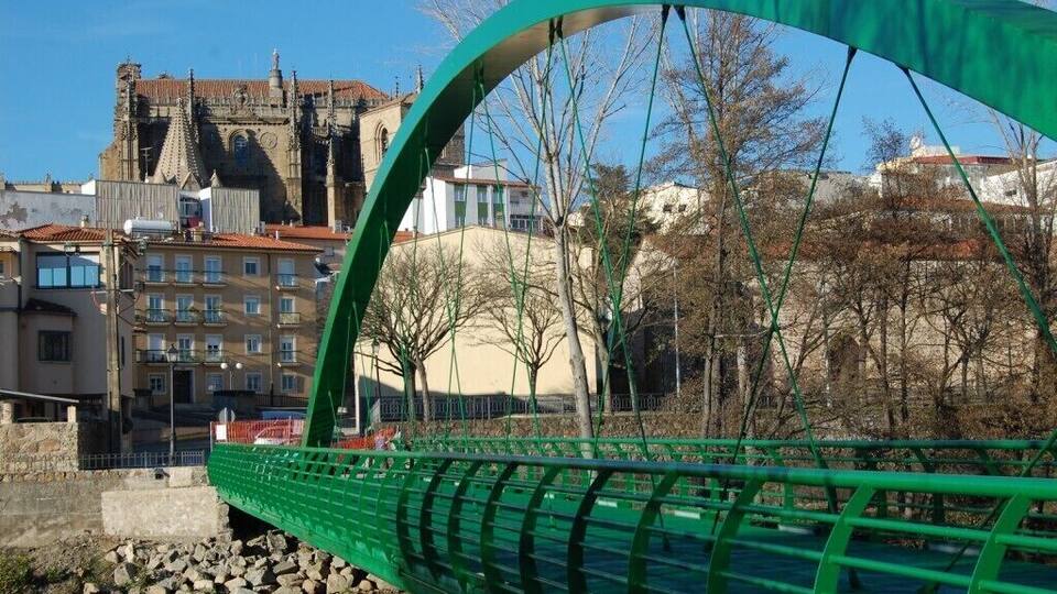 A green pedestrian bridge with a curved arch, a stone embankment, and buildings in the background.