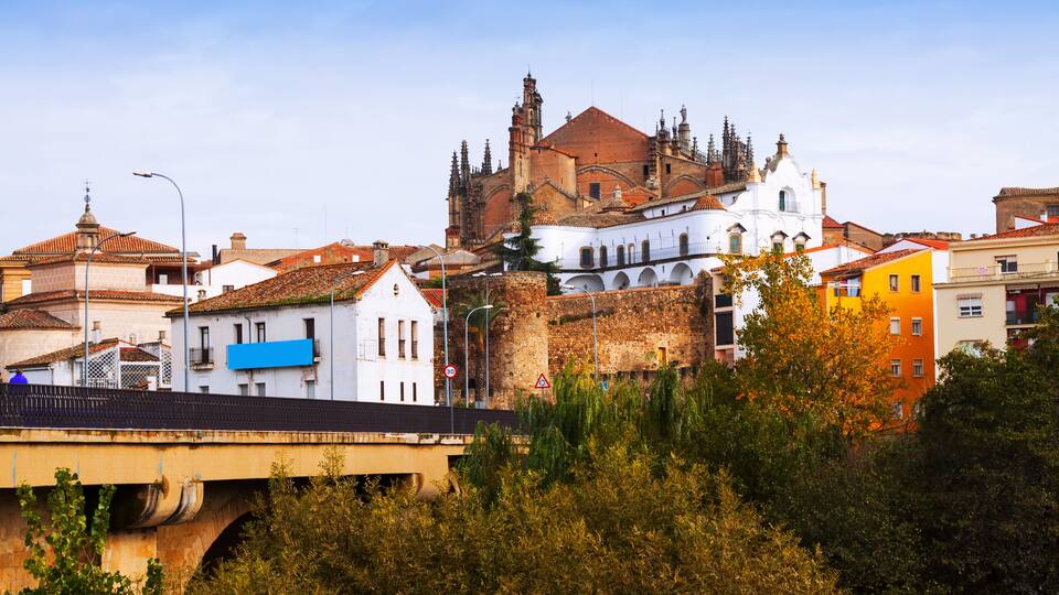 A historic cathedral with a red roof and white walls, surrounded by colorful buildings and a bridge.
