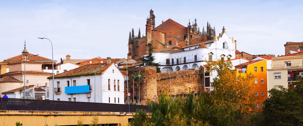 A historic cathedral with a red roof and white walls, surrounded by colorful buildings and a bridge.
