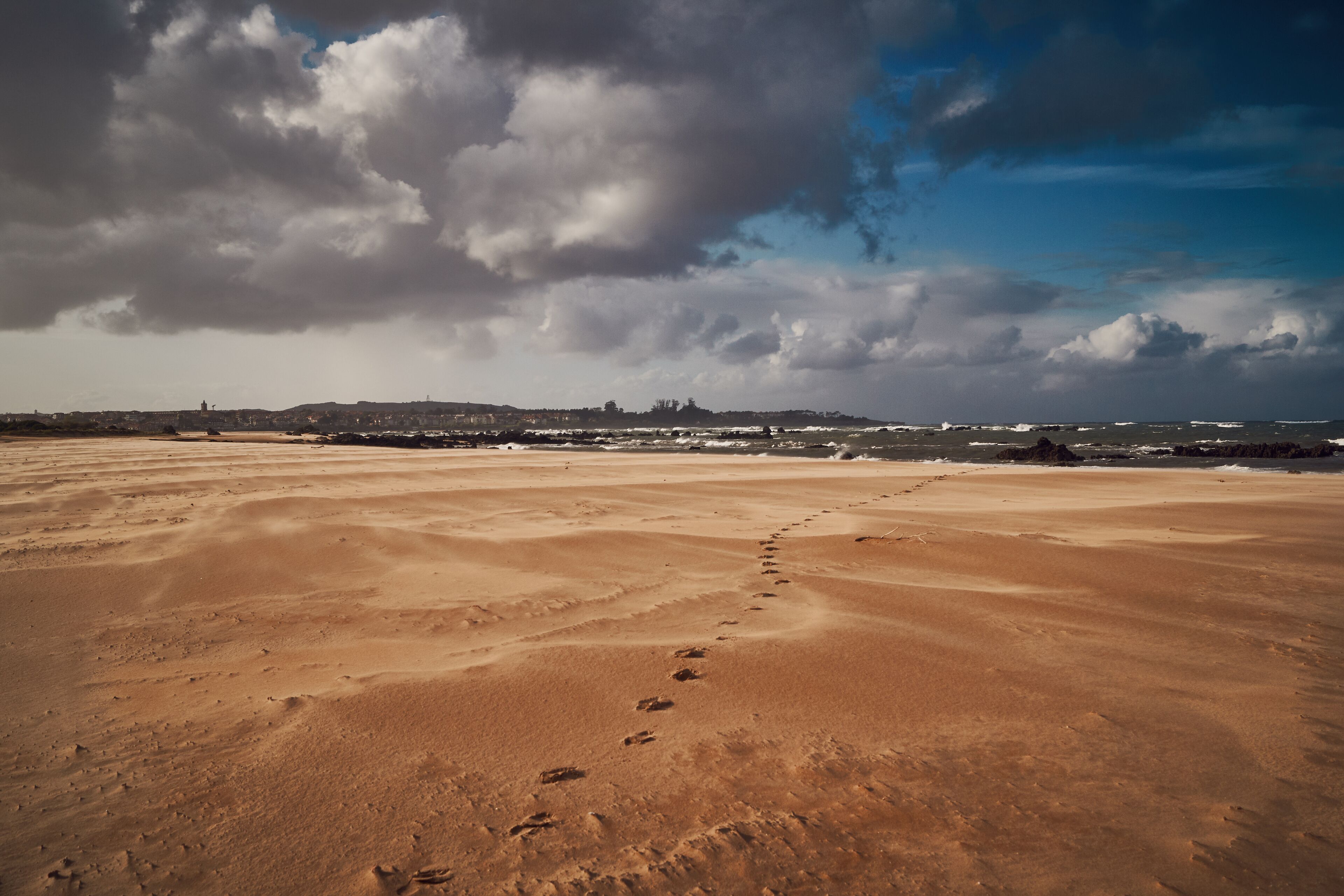 A desert landscape with footprints leading towards a distant city skyline under a cloudy sky.