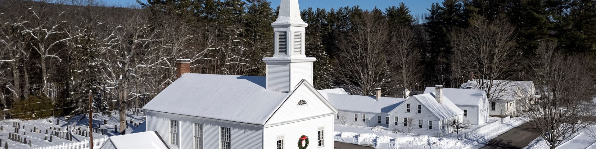 A white church with a steeple, surrounded by snow-covered trees and a clear blue sky.