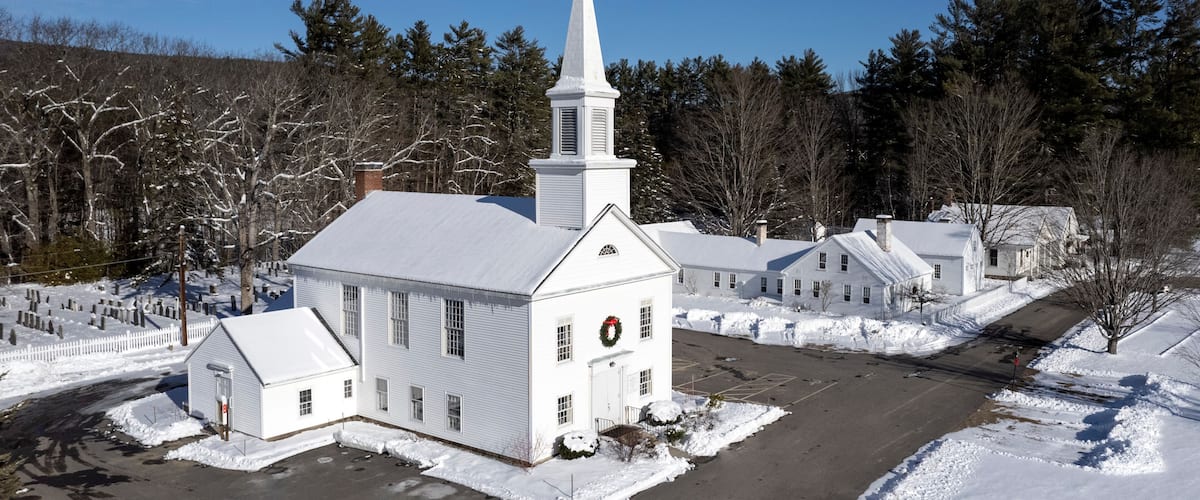A white church with a steeple, surrounded by snow-covered trees and a clear blue sky.