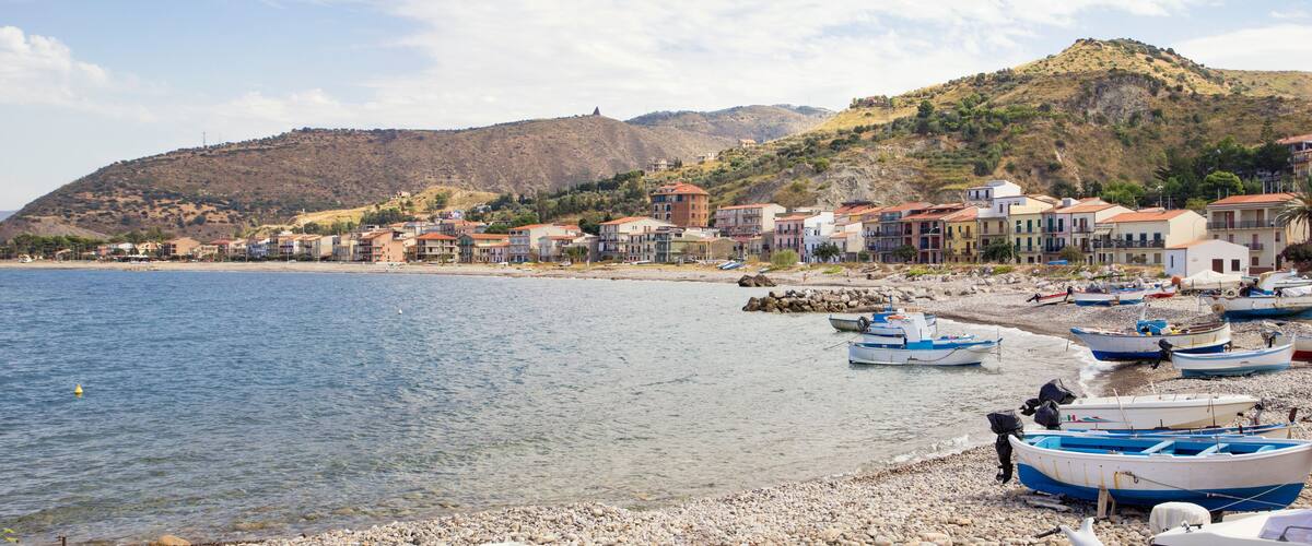A coastal town with buildings along the shore, boats docked on the beach, and hills in the background.