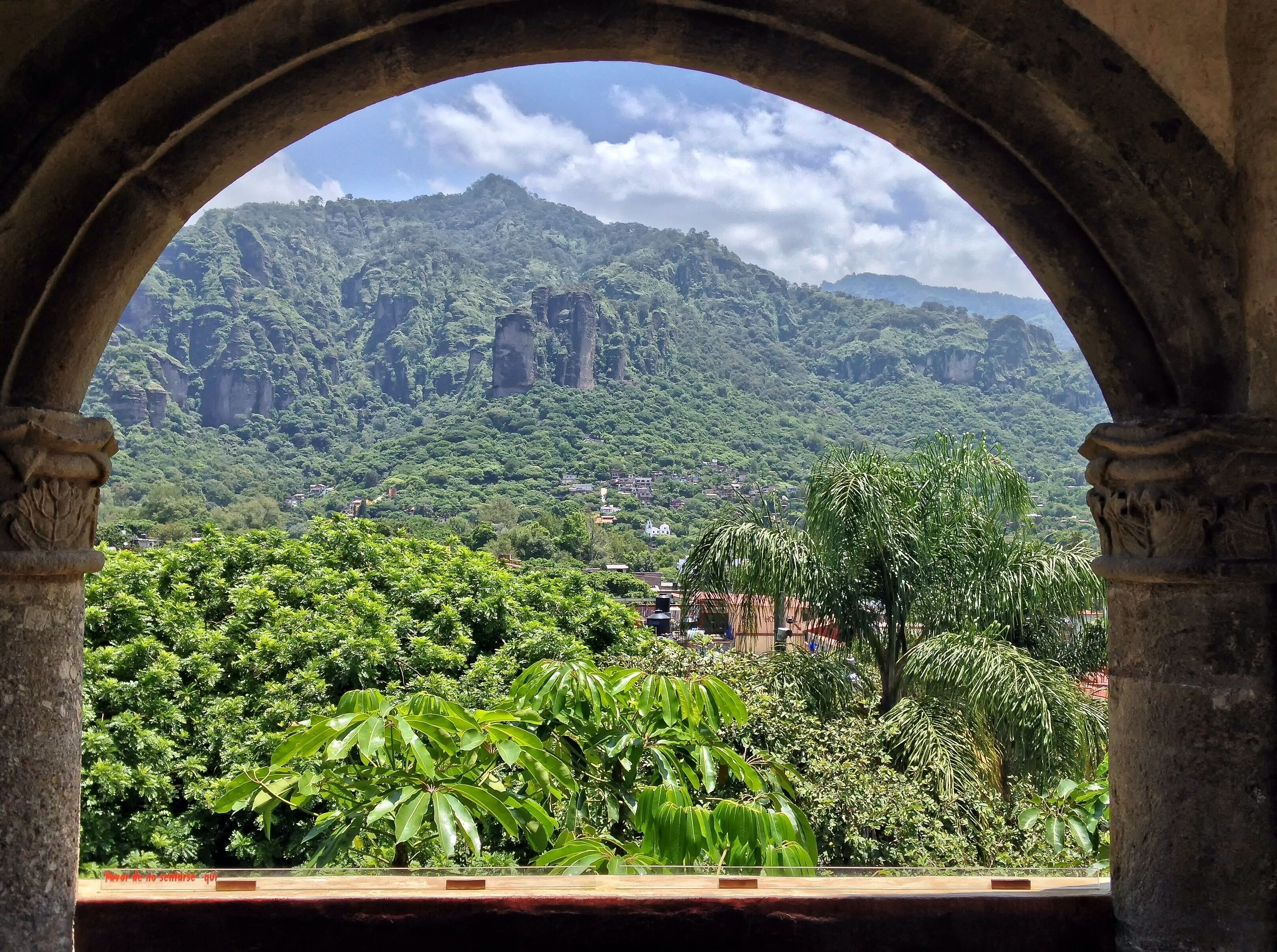 Vista a través de un arco de un paisaje verde exuberante con abundante follaje y una cadena montañosa distante.