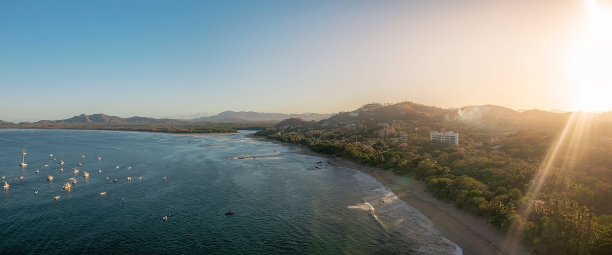 A coastal landscape with a beach, boats, and a hilly area with buildings.