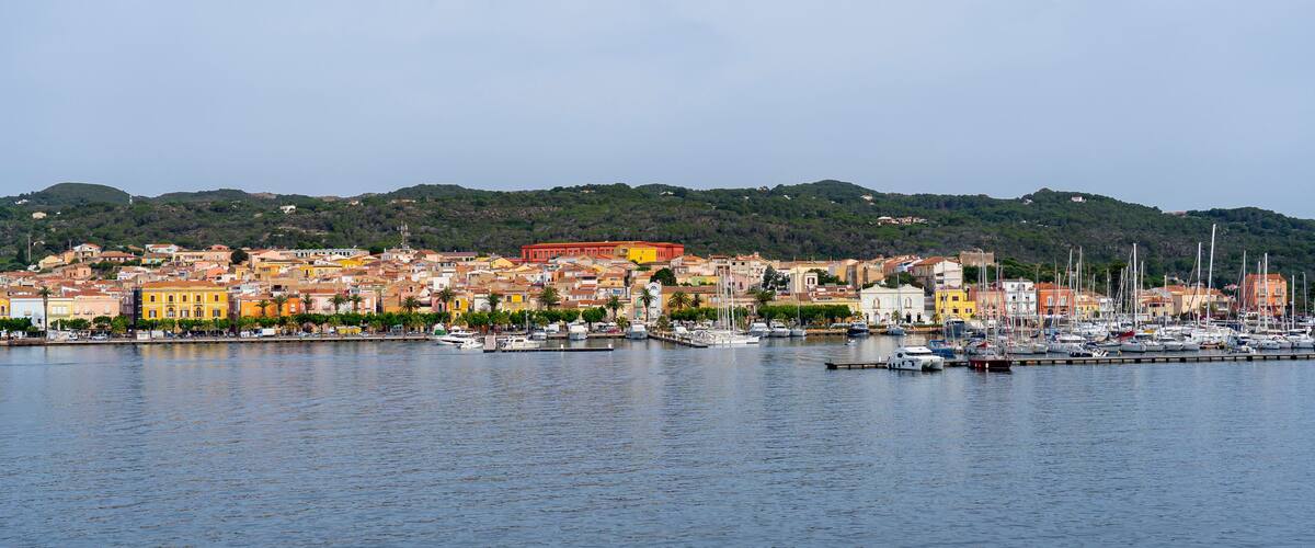 A coastal town with colorful buildings, a marina with boats, and hills in the background.