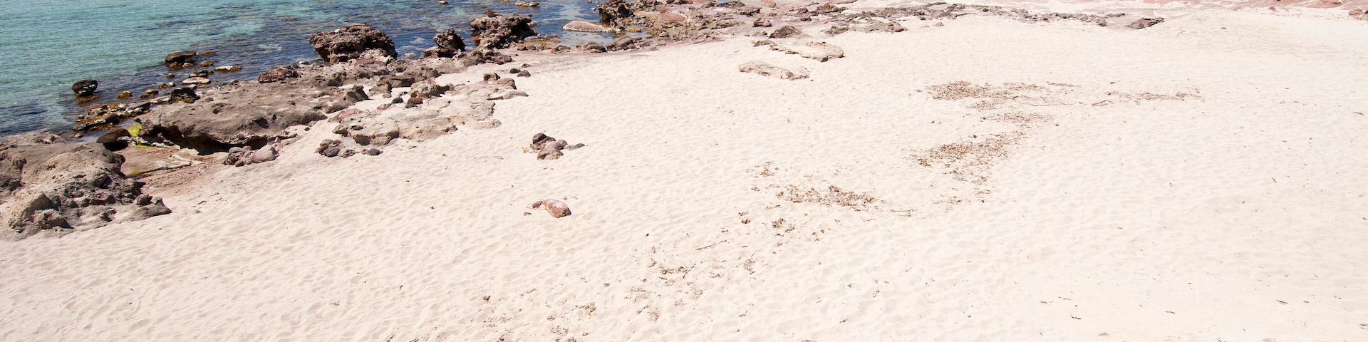 Una playa arenosa con agua turquesa cristalina, afloramientos rocosos y una colina verde al fondo.