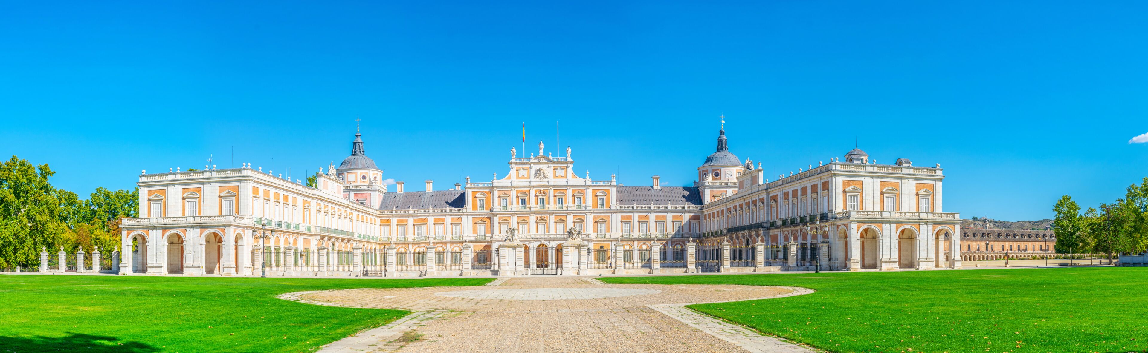 A large, historic building with a central dome and two smaller domes, surrounded by a well-maintained lawn and a gravel pathway leading to the entrance.