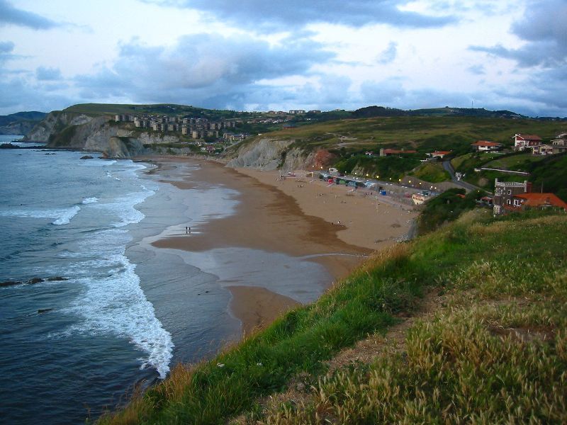 Un paisaje costero con una playa arenosa, acantilados y un pueblo al fondo.