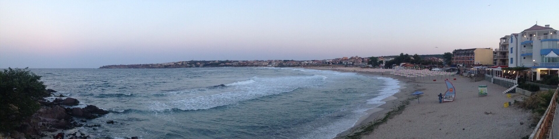 Uma praia com ondas, areal arenoso e edifícios ao fundo.