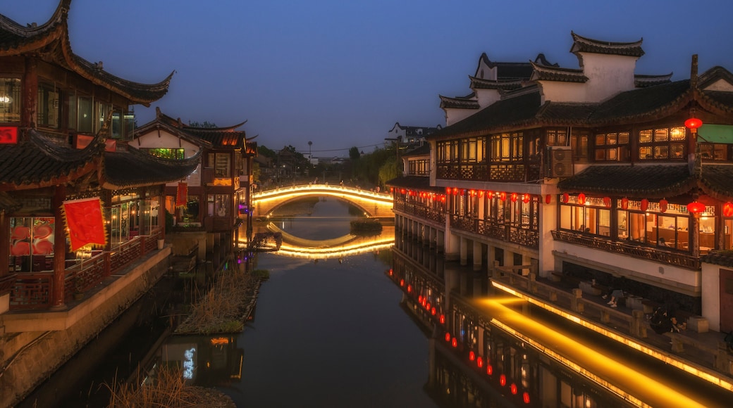 Un puente chino tradicional sobre un canal al anochecer, con edificios iluminados a ambos lados.