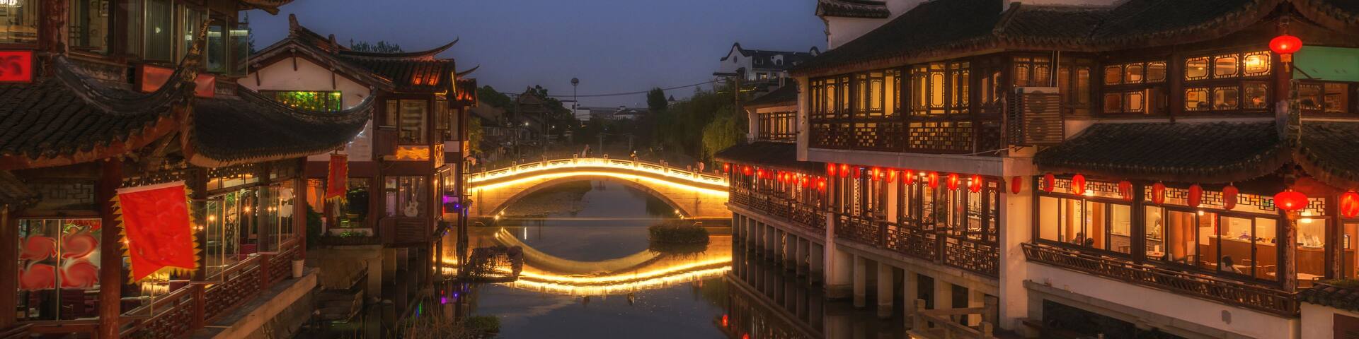 A traditional Chinese bridge over a canal at dusk, with illuminated buildings on either side.