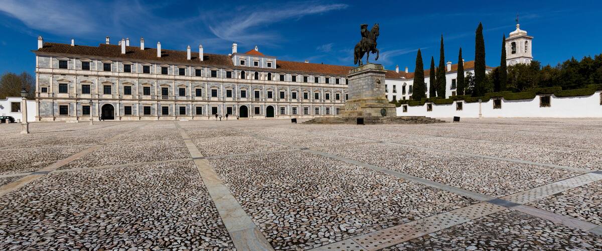 A historic building with a courtyard paved with pebbles, a statue, and a bell tower.