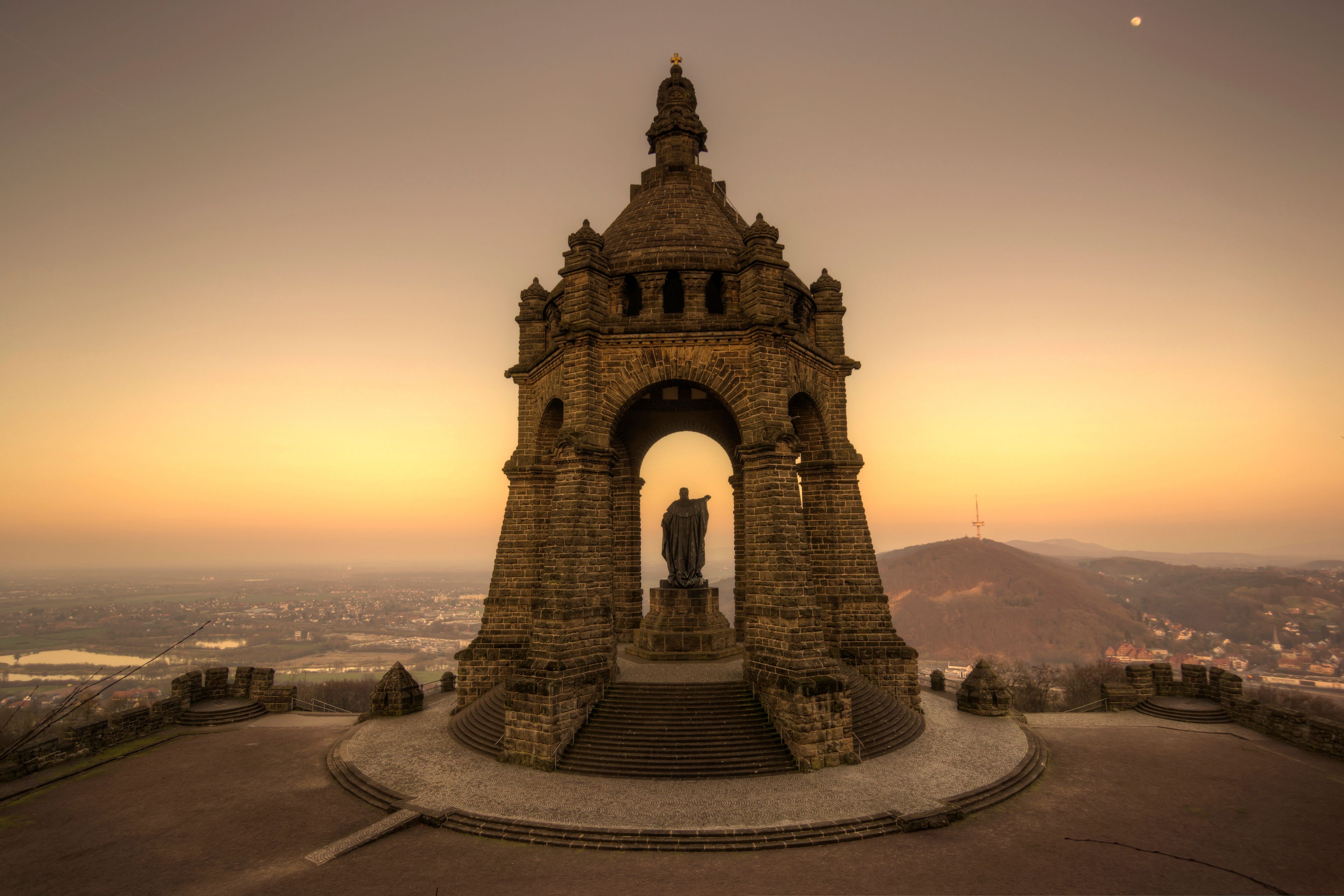 Un arc en pierre d’époque, surmonté d’une statue, est entouré d’une plateforme circulaire en pierre et accessible par des marches.