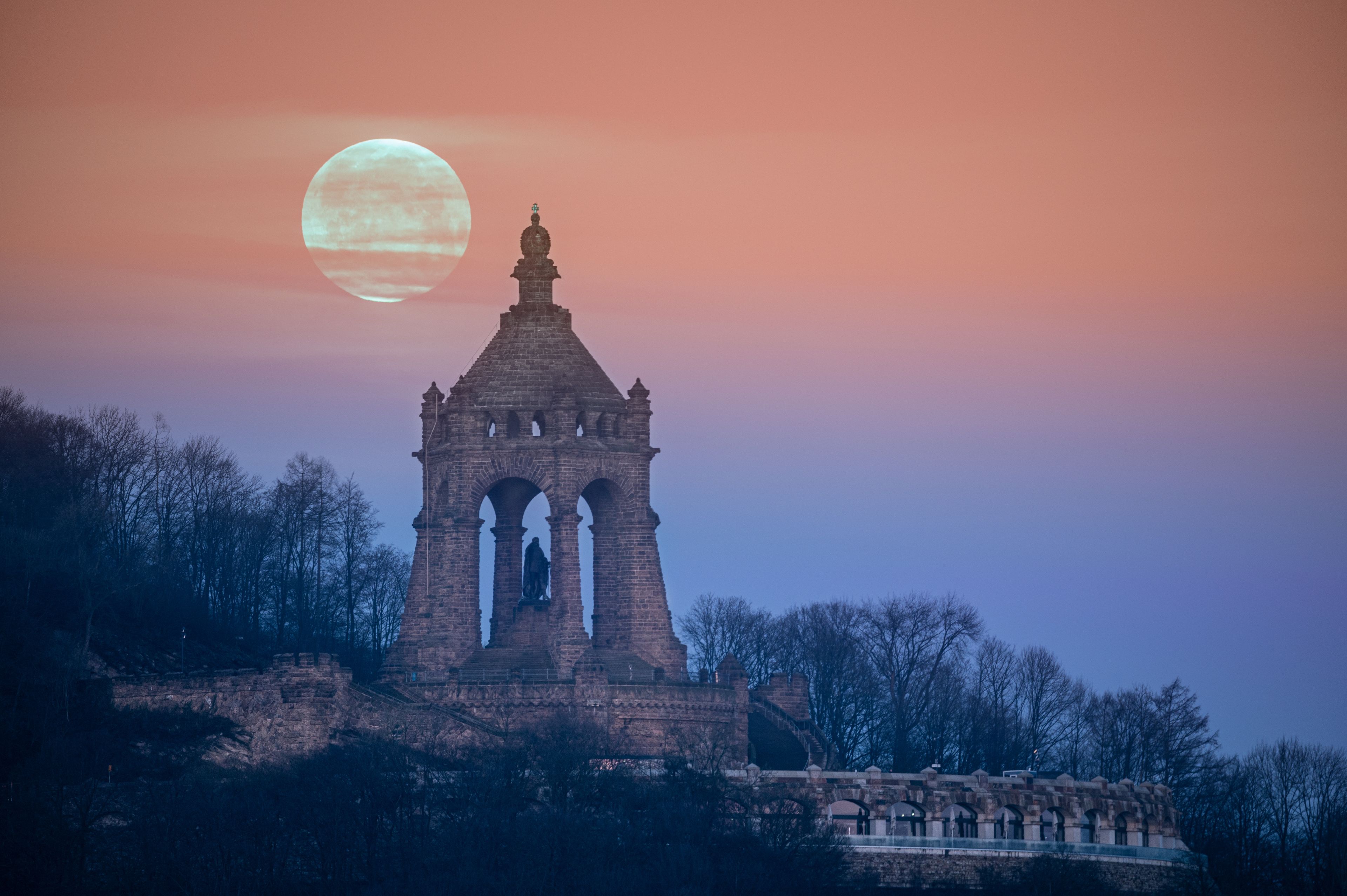 Une tour historique, ornée d’arches et d’une statue, se dressant sur fond de ciel crépusculaire et d’une grande lune.