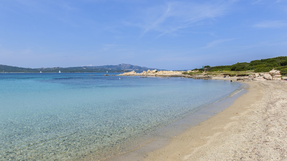 A clear beach with shallow water, rocky shoreline, and distant mountains.