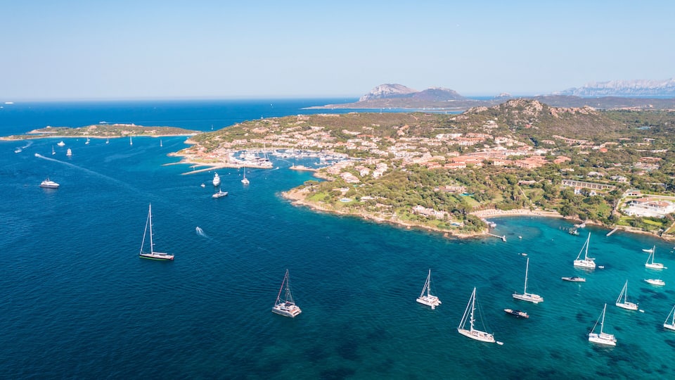 A coastal town with sailboats in the water, surrounded by hills and clear blue sea.