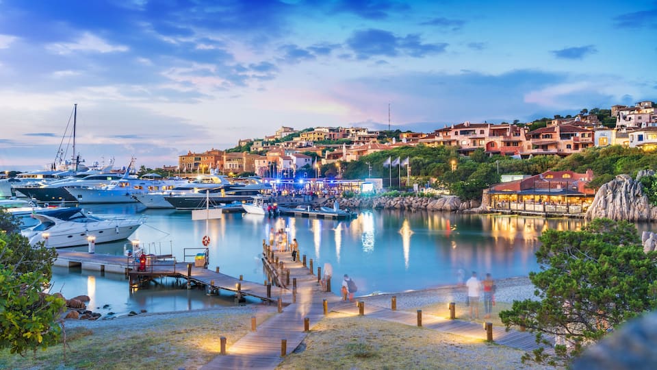 A marina with yachts, a wooden dock, and a hillside town with buildings.