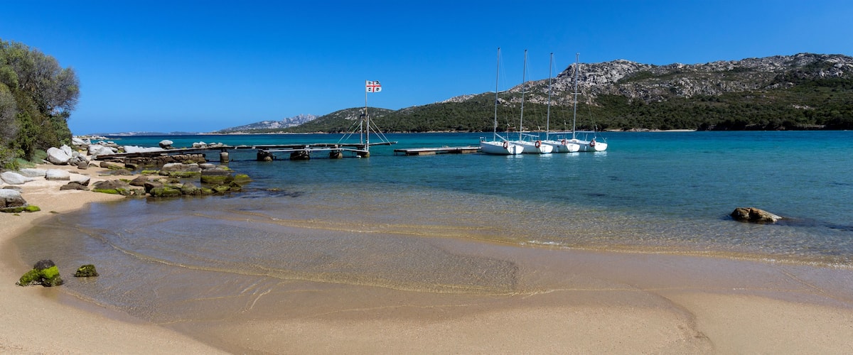 A clear turquoise sea, a sandy beach, and rocky outcrops with boats docked at a pier.