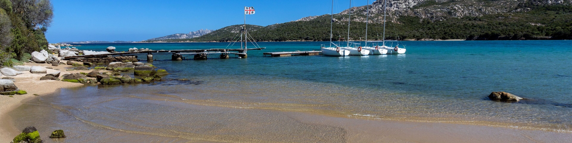 A clear turquoise sea, a sandy beach, and rocky outcrops with boats docked at a pier.