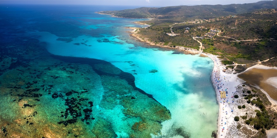 Vue aérienne d’une zone côtière avec des eaux turquoise limpides, une plage de sable et un paysage vallonné en arrière-plan.