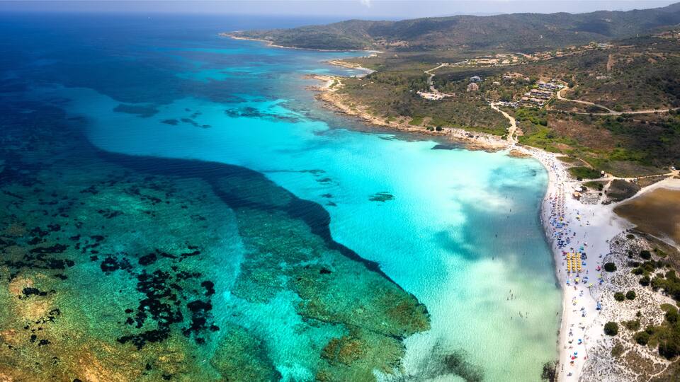 An aerial view of a coastal area with clear turquoise waters, a sandy beach, and a hilly landscape in the background.