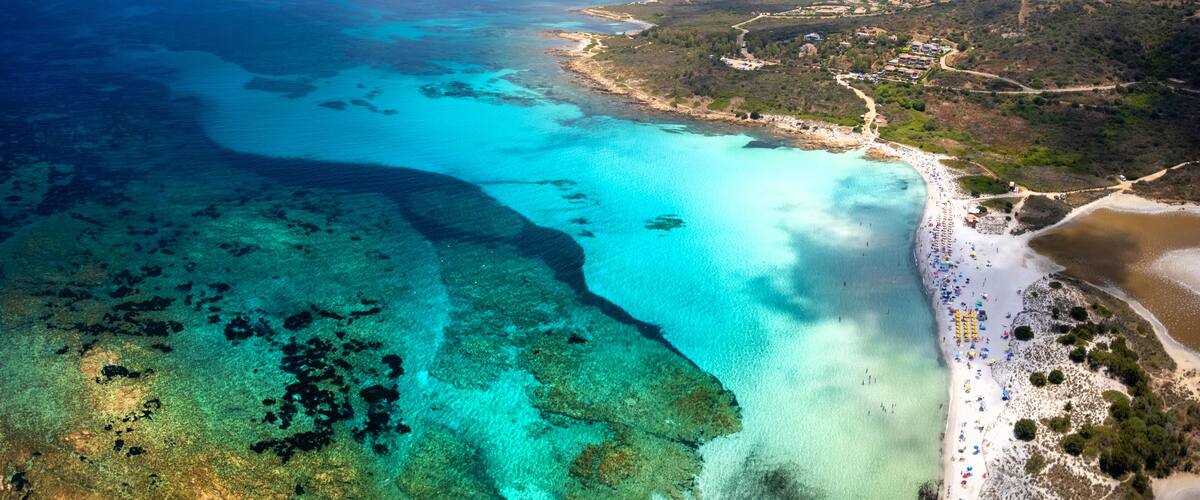 An aerial view of a coastal area with clear turquoise waters, a sandy beach, and a hilly landscape in the background.