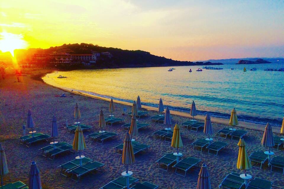 A beach with rows of sun loungers and umbrellas, a calm sea, and a sunset in the background.