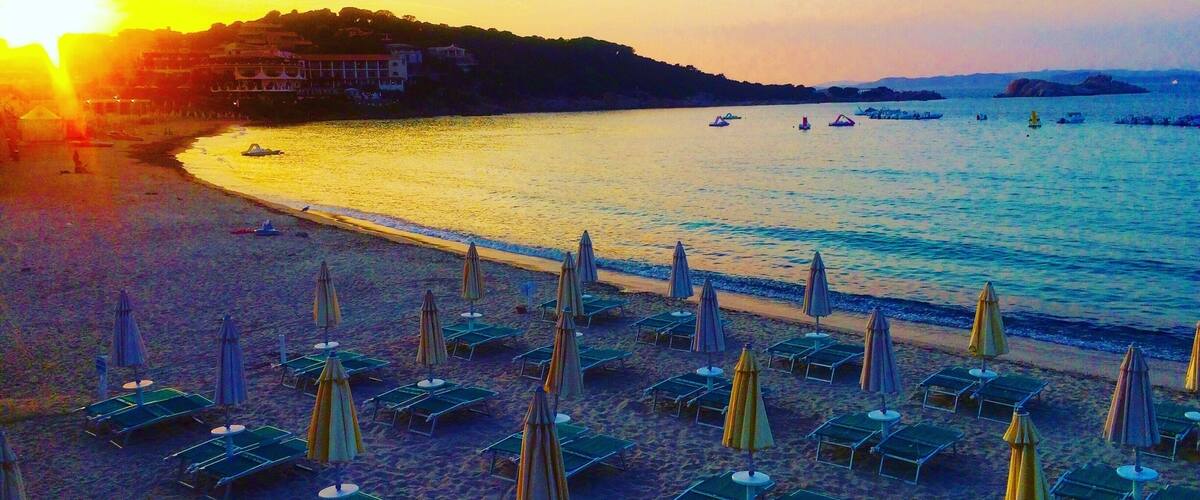 A beach with rows of sun loungers and umbrellas, a calm sea, and a sunset in the background.